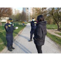 zdjęcie przedstawia strażników miejskich informujących kobietę o nakazach i zakazach wprowadzonych przez rząd. Strażnicy mają na twarzach maseczki, kobieta ma nos i usta zasłonięte apaszką.