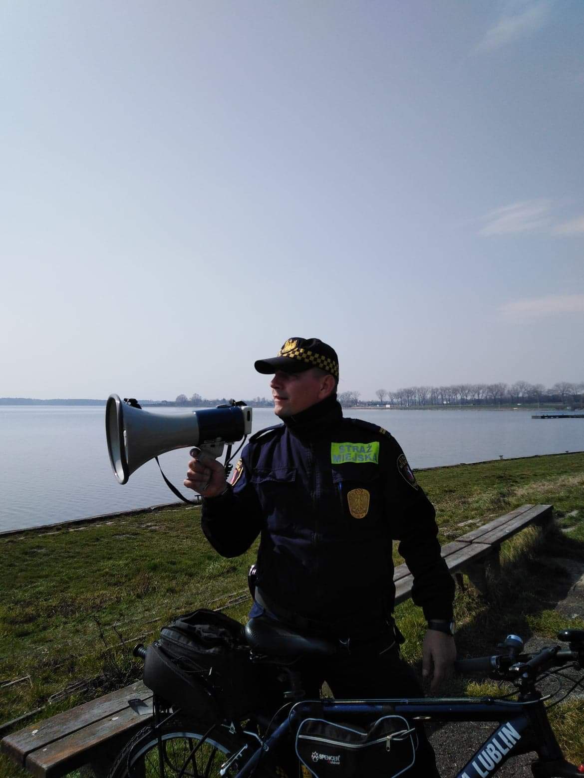 Zdjęcie przedstawia strażnika miejskiego z megafonem na tle Zalewu Zemborzyckiego w Lublinie