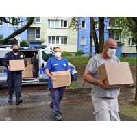 Strażnik miejski oraz dwóch pracowników szkoły niosą pudełka z termometrami i maseczkami